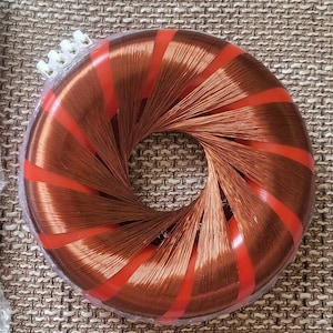 Puede incluir: Un inductor toroidal de color cobre con tiras de plástico rojo. El inductor está envuelto en plástico transparente y tiene un conector blanco en el lateral. Los cables están enrollados alrededor de un núcleo circular.