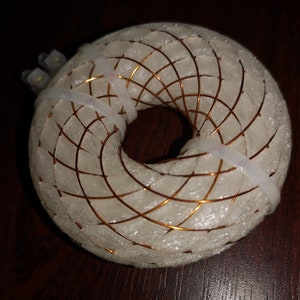 May include: A white, donut-shaped object with a copper wire grid pattern. The object is wrapped with clear plastic ties.