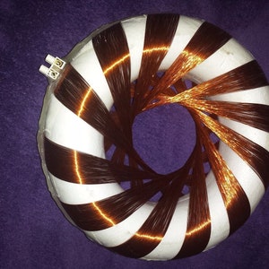 May include: A toroidal inductor with a white core and alternating bands of dark brown and white. Copper wire is wound around the core, creating a circular pattern. Two white connectors are visible on the top. The object is set against a purple background.