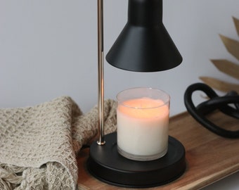 May include: A black electric candle warmer with a gold metal arm and a black shade. A white candle is placed on the warmer's base. The warmer is sitting on a wooden tray.