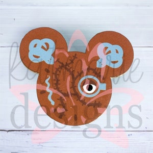 May include: A brown Mickey Mouse-shaped pin with a stitched, blue-eyed monster face and blue stitching details.