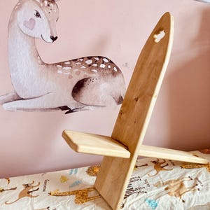 May include: A small, light-colored wooden chair with a heart-shaped cutout at the top. The chair is positioned in front of a pink wall decorated with a deer and an owl illustration. The chair is designed for children.
