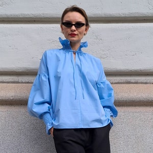 K&ouml;nnte beinhalten: Hellblaue Bluse mit R&uuml;schenkragen und weiten &Auml;rmeln. Die Bluse wird mit einer schwarzen Hose mit weitem Bein und schwarzen Riemchen-High Heels kombiniert. Das Outfit befindet sich vor einer hellgrauen Wand.