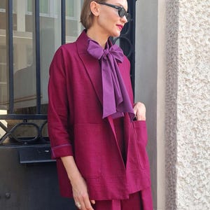 May include: A magenta blazer with three-quarter length sleeves, two front patch pockets, and a notched lapel, paired with a matching magenta bottom and a large, dark purple pussy-bow blouse worn underneath with a visible bow at the neckline.