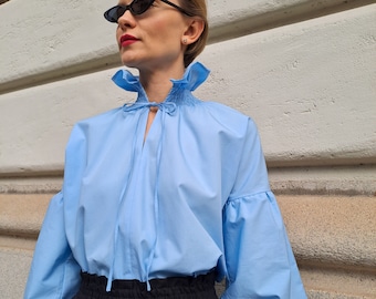 Könnte beinhalten: Hellblaue Bluse mit gerüschtem Kragen und langen, puffärmeligen Ärmeln. Die Bluse hat eine Schleife am Ausschnitt und wird mit einer schwarzen Hose kombiniert. Die Ärmel haben elastische Bündchen. Der Stoff ist leicht.