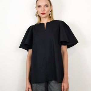 May include: A black top with short, flared sleeves and a notched neckline. The top is made of a solid, opaque fabric. The model is wearing gold earrings and gray pants.