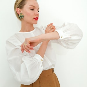Puede incluir: Una blusa blanca con mangas largas y abullonadas y pu&ntilde;os con botones dorados. La modelo lleva anillos dorados y pendientes verdes y dorados. La modelo tambi&eacute;n lleva unos pantalones cortos marrones.
