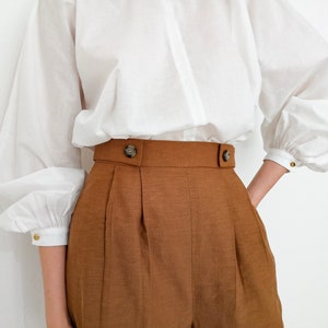 Puede incluir: Una blusa blanca con mangas voluminosas y cuello alto combinada con pantalones cortos marrones de cintura alta. Los pantalones cortos tienen una cintura ancha con dos botones decorativos. Las mangas tienen pu&ntilde;os abotonados.