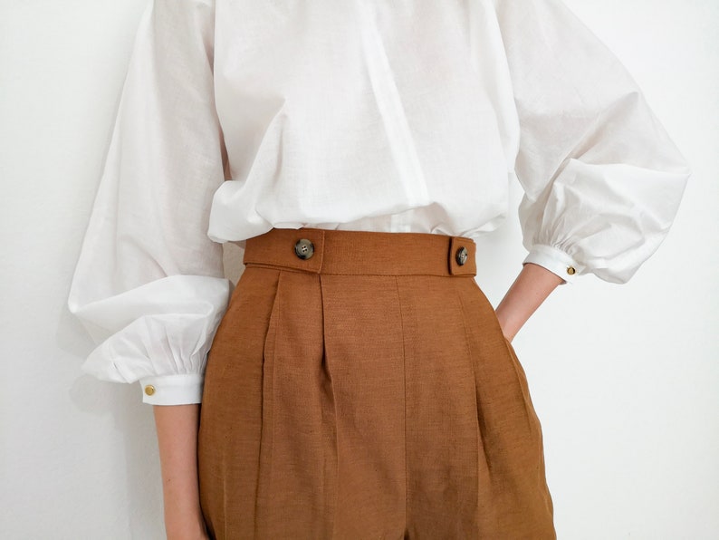 Puede incluir: Una blusa blanca con mangas voluminosas y cuello alto combinada con pantalones cortos marrones de cintura alta. Los pantalones cortos tienen una cintura ancha con dos botones decorativos. Las mangas tienen pu&ntilde;os abotonados.
