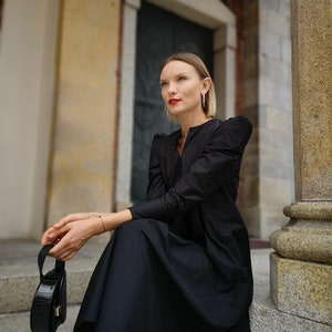 May include: A woman wearing a black dress with long sleeves and a round black handbag. The dress has a V-neck and a flowing skirt. The woman is wearing gold hoop earrings and red lipstick. She is sitting on stone steps.
