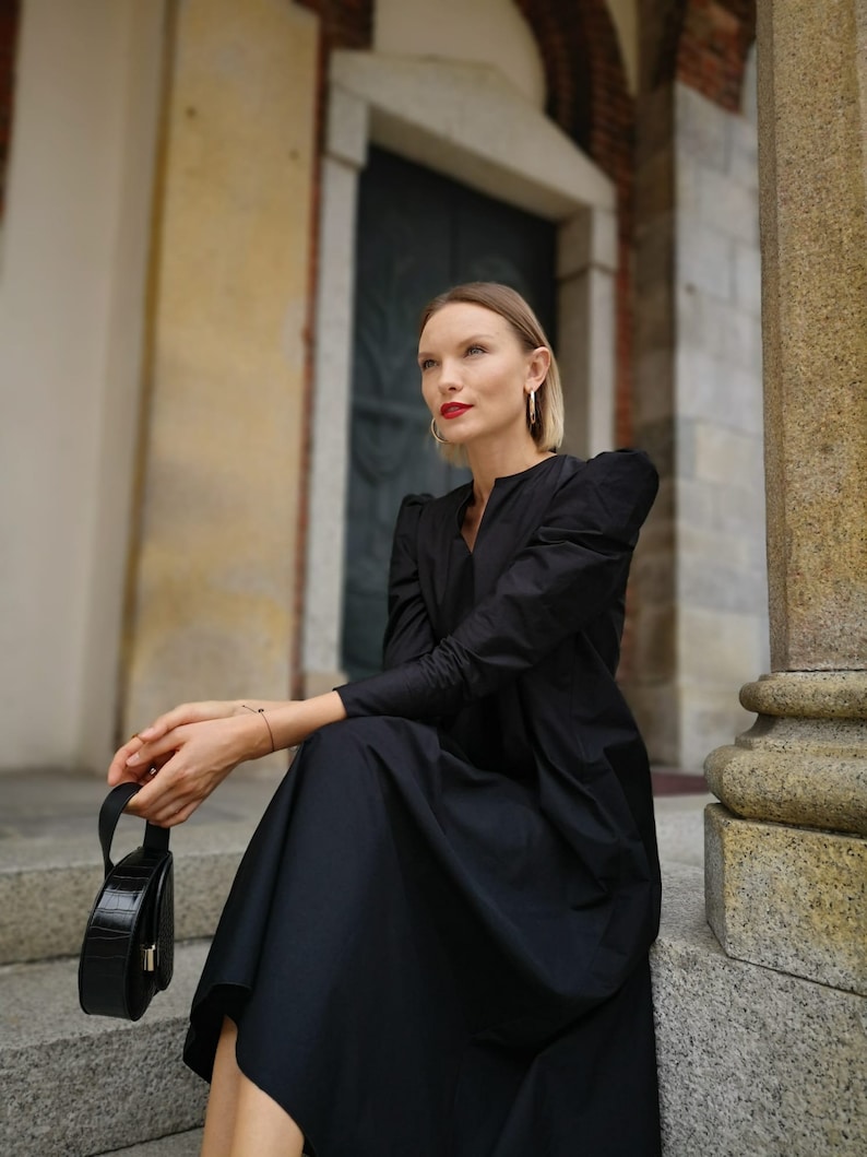 May include: A woman wearing a black dress with long sleeves and a round black handbag. The dress has a V-neck and a flowing skirt. The woman is wearing gold hoop earrings and red lipstick. She is sitting on stone steps.