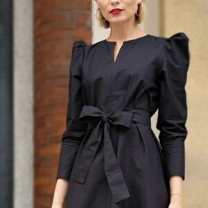 May include: Black dress with a notched neckline, long sleeves, and a bow-tie waist. The dress features puffed shoulders and a knee-length skirt. The fabric appears to be a smooth, woven material. The dress is a solid black color.
