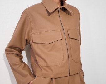 Könnte beinhalten: Eine braune Reißverschlussjacke mit Kragen und zwei großen Taschen. Die Jacke wird mit einer passenden braunen Hose kombiniert.