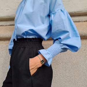 K&ouml;nnte beinhalten: Eine hellblaue Bluse mit volumin&ouml;sen &Auml;rmeln und ger&uuml;schten Manschetten wird mit einer schwarzen Hose kombiniert. Eine goldene Uhr mit quadratischem Zifferblatt ist sichtbar. Das Outfit ist schlicht und stilvoll.
