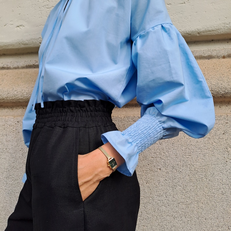 K&ouml;nnte beinhalten: Eine hellblaue Bluse mit volumin&ouml;sen &Auml;rmeln und ger&uuml;schten Manschetten wird mit einer schwarzen Hose kombiniert. Eine goldene Uhr mit quadratischem Zifferblatt ist sichtbar. Das Outfit ist schlicht und stilvoll.