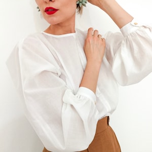 Puede incluir: Una blusa blanca con mangas voluminosas y detalles de botones dorados. La blusa tiene un cuello redondo y se combina con pantalones marrones. Las mangas est&aacute;n recogidas en los pu&ntilde;os. La modelo lleva pintalabios rojo.