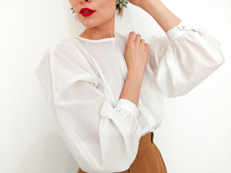 Puede incluir: Una blusa blanca con mangas voluminosas y detalles de botones dorados. La blusa tiene un cuello redondo y se combina con pantalones marrones. Las mangas est&aacute;n recogidas en los pu&ntilde;os. La modelo lleva pintalabios rojo.