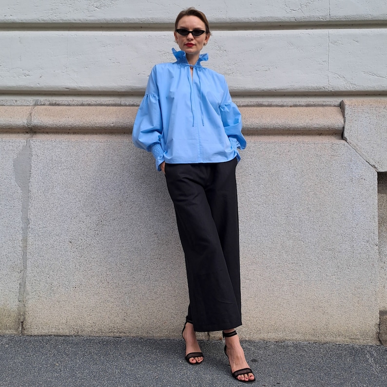 K&ouml;nnte beinhalten: Hellblaue Bluse mit R&uuml;schenkragen und weiten &Auml;rmeln. Die Bluse wird mit einer schwarzen Hose mit weitem Bein und schwarzen Riemchen-High Heels kombiniert. Das Outfit befindet sich vor einer hellgrauen Wand.