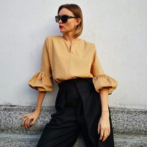 May include: A tan-colored blouse with three-quarter length, puffy sleeves and a V-neckline. The blouse is paired with high-waisted, wide-leg black pants. The person is wearing black sunglasses.