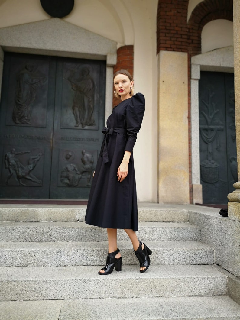 May include: A black midi dress with long sleeves and a tie waist. The dress features puffed sleeves and is paired with black open-toe heels. The background includes a stone staircase and a large, dark door.