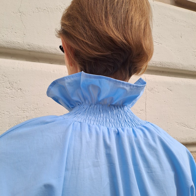 K&ouml;nnte beinhalten: Eine hellblaue Bluse mit einem hohen, ger&uuml;schten Kragen. Der Kragen ist am Halsausschnitt gerafft, wodurch ein strukturierter Effekt entsteht. Die Bluse ist aus einem glatten, leichten Stoff gefertigt.
