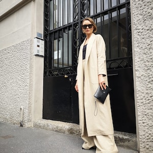 Puede incluir: Una mujer con un largo abrigo de lino beige y pantalones anchos a juego. Está de pie frente a una puerta de metal negro con un bolso de mano negro en la mano.