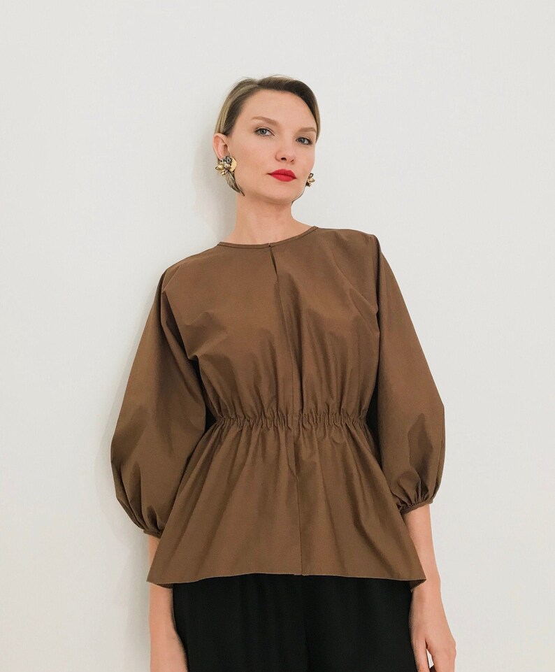 May include: A brown blouse with three-quarter sleeves and an elastic waist. The top has a round neckline and a peplum-style hem. The model is wearing black pants and gold earrings.