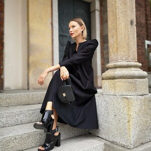 May include: A woman wearing a black dress, black open-toe heels, and carrying a black handbag with a gold clasp. The dress has puffed sleeves. The woman is sitting on stone steps next to a large column.