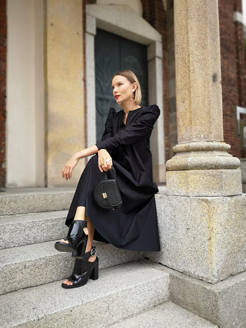 May include: A woman wearing a black dress, black open-toe heels, and carrying a black handbag with a gold clasp. The dress has puffed sleeves. The woman is sitting on stone steps next to a large column.