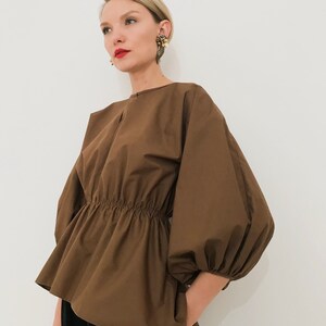 May include: A brown top with a gathered waist and voluminous sleeves. The top has a round neckline and a peplum-style hem. The sleeves are gathered at the wrist. The model is wearing black pants.