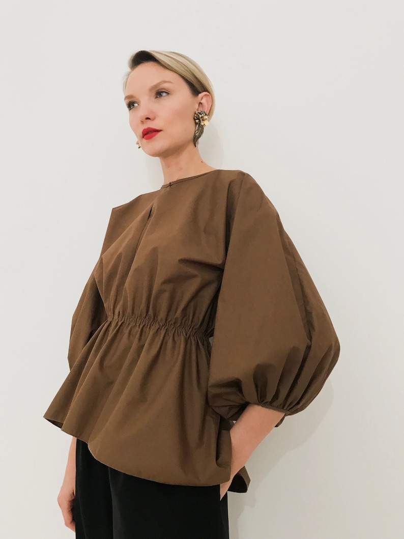 May include: A brown top with a gathered waist and voluminous sleeves. The top has a round neckline and a peplum-style hem. The sleeves are gathered at the wrist. The model is wearing black pants.