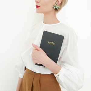 Puede incluir: Un cuaderno negro con la palabra dorada "NOTES" se sostiene frente a una blusa blanca. El cuaderno se sostiene en la mano. La persona lleva una blusa blanca y pantalones marrones. Los pendientes son verdes y dorados.