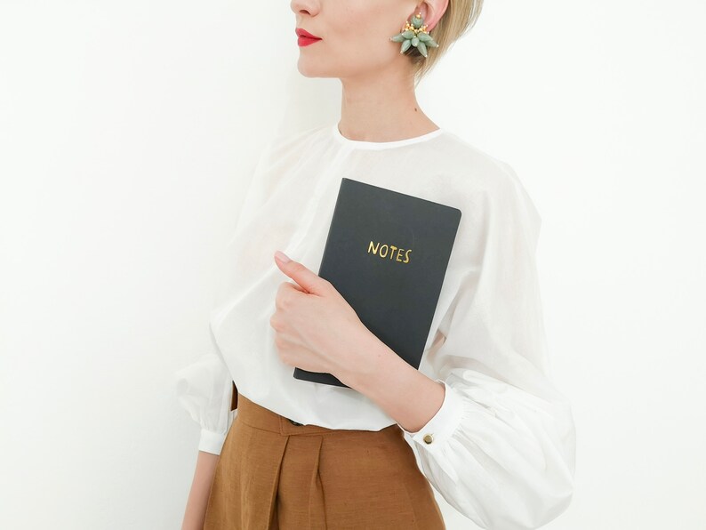 Puede incluir: Un cuaderno negro con la palabra dorada "NOTES" se sostiene frente a una blusa blanca. El cuaderno se sostiene en la mano. La persona lleva una blusa blanca y pantalones marrones. Los pendientes son verdes y dorados.