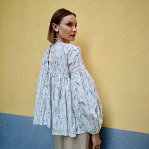 May include: A white blouse adorned with a pattern of green and blue leaves. It features long, voluminous sleeves and a gathered waist. The back has a tie closure at the neckline. The background is a yellow wall with a blue stripe.