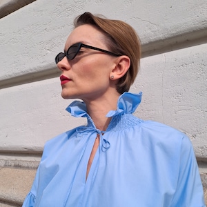 K&ouml;nnte beinhalten: Hellblaue Bluse mit R&uuml;schenkragen und Bindedetail. Die Bluse hat lange &Auml;rmel und wird mit schwarzen Cat-Eye-Sonnenbrillen kombiniert. Die Person tr&auml;gt einen kleinen Ohrstecker.