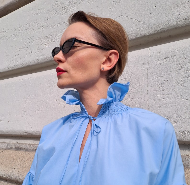 K&ouml;nnte beinhalten: Hellblaue Bluse mit R&uuml;schenkragen und Bindedetail. Die Bluse hat lange &Auml;rmel und wird mit schwarzen Cat-Eye-Sonnenbrillen kombiniert. Die Person tr&auml;gt einen kleinen Ohrstecker.