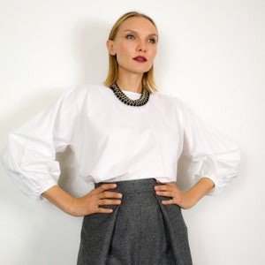 May include: A woman wearing a white long-sleeve blouse with puffed sleeves and gray high-waisted pants. She is wearing a black and gold beaded necklace. The background is a plain white wall.