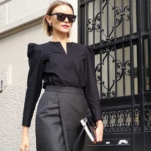 May include: A woman wearing a black long-sleeve blouse with puffed sleeves and a gray wrap skirt. She is wearing black sunglasses and holding a black clutch. The background includes a white wall and a black metal gate.