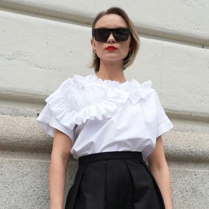 May include: A white ruffled blouse with short sleeves and a black skirt. The woman is wearing black sunglasses and red lipstick.
