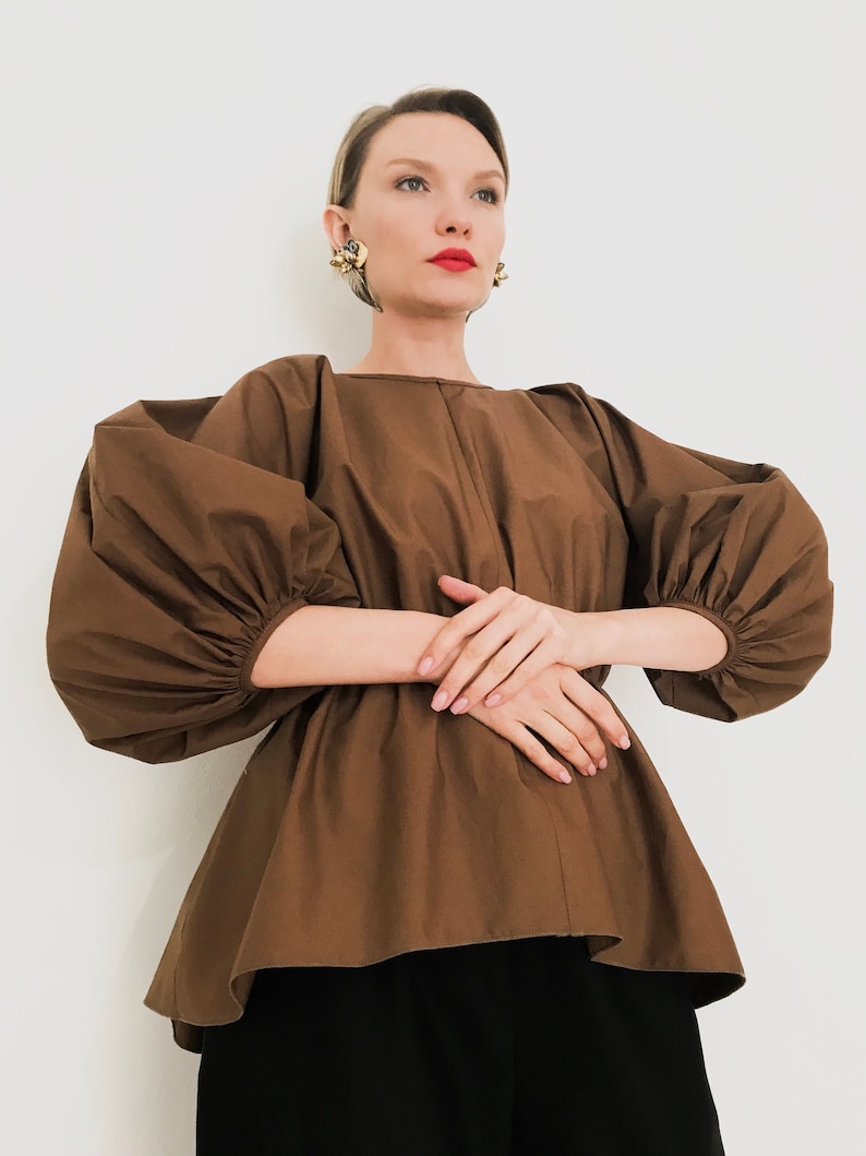 May include: A brown blouse with voluminous, puffed sleeves and a peplum hem. The top is paired with black pants. The model is wearing gold earrings and red lipstick. The blouse is made of a smooth, solid-colored fabric.
