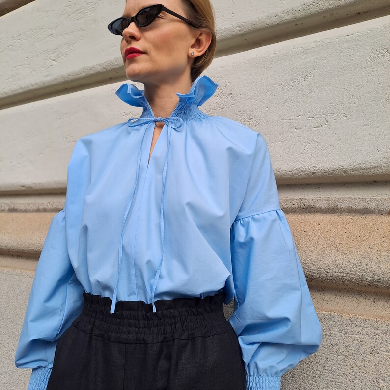 K&ouml;nnte beinhalten: Hellblaue Bluse mit ger&uuml;schtem Kragen und langen, puff&auml;rmeligen &Auml;rmeln. Die Bluse hat eine Schleife am Ausschnitt und wird mit einer schwarzen Hose kombiniert. Die &Auml;rmel haben elastische B&uuml;ndchen. Der Stoff ist leicht.