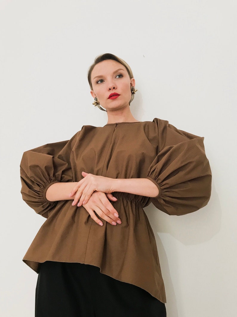 May include: A brown blouse with voluminous, gathered sleeves and a cinched waist. The top has a unique asymmetrical hemline and is paired with black bottoms. The model is wearing gold earrings and red lipstick.