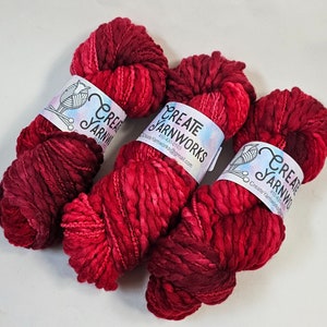 Pode incluir: Tr&ecirc;s novelos de fio tingido &agrave; m&atilde;o em vermelho escuro e bord&ocirc; com um r&oacute;tulo branco que diz "Create Yarnworks" e um endere&ccedil;o de site.