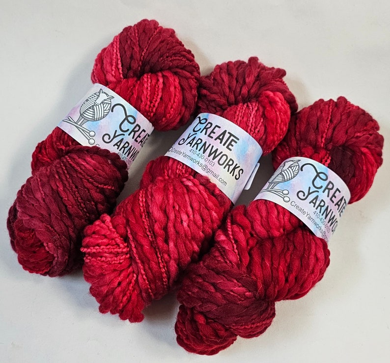 Pode incluir: Tr&ecirc;s novelos de fio tingido &agrave; m&atilde;o em vermelho escuro e bord&ocirc; com um r&oacute;tulo branco que diz "Create Yarnworks" e um endere&ccedil;o de site.