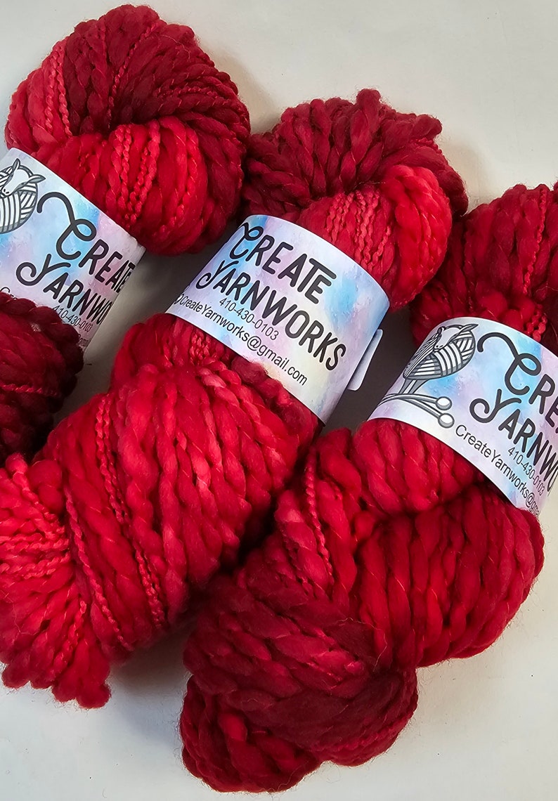 Pode incluir: Tr&ecirc;s novelos de fio vermelho e rosa com um r&oacute;tulo branco que diz "Create Yarnworks 410-430-0103 CreateYarnworks@gmail.com"