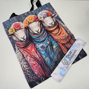 May include: Three sheep wearing colorful floral headbands and scarves. The sheep are standing side-by-side and looking forward. The background is a dark blue with a floral pattern. The tote bag is made of canvas and has black handles. The bag has the text "Create Yarnworks" on the bottom right corner.