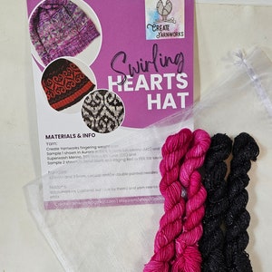 May include: A kit for knitting a hat with a swirling hearts pattern. The kit includes two skeins of yarn, one black and one pink, and a printed pattern card with instructions. The card includes the text "Swirling Hearts Hat" and "Create Yarnworks".