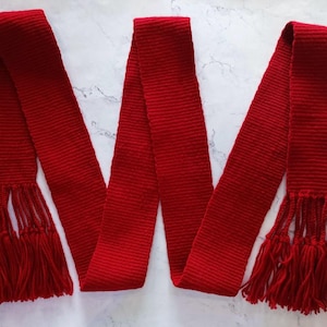 May include: A vibrant red scarf with a ribbed texture and fringed ends. The scarf is made of a soft material and is designed to be worn around the neck. The scarf is approximately 72 inches long.