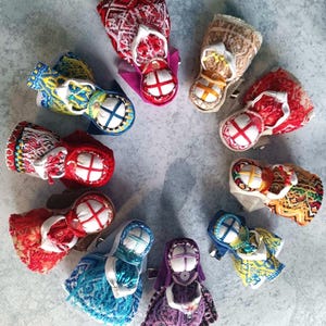 Puede incluir: Una disposición circular de diez pequeñas muñecas de tela hechas a mano. Cada muñeca presenta una cabeza blanca con un patrón de cruz y viste ropa colorida con bordados intrincados en tonos rojo, azul, amarillo, morado y beige.