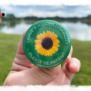 May include: A green button featuring a yellow sunflower and the text "I HAVE A HIDDEN DISABILITY PLEASE BE PATIENT". The button is held in a hand, with a blurred outdoor scene in the background.